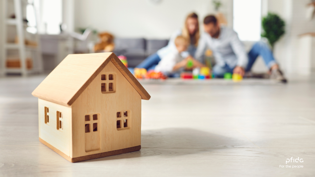 A wooden toy house with a family blurred in the background | 3 Key Insights from Pfida's Shariah-Compliant Home Finance Podcast with Sheikh Asim Khan